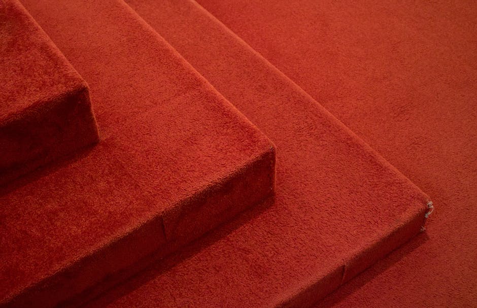 Close-up view of a deep red carpeted staircase in a residential setting, showcasing clean, plush carpet fibers with no visible dirt or stains. The stairs are made of a soft-textured material, possibly wool or synthetic fiber, and are evenly carpeted with no loose edges or damage. The top step is slightly taller, indicating a standard staircase design, with lighting that highlights the uniformity and cleanliness of the surface. The surrounding area appears well-maintained, reflecting effective surface cleaning and deep cleaning processes typical of Roehampton Carpet Cleaning's domestic cleaning services. This image emphasizes the importance of regular hygiene and maintained carpet surfaces for a tidy home environment.