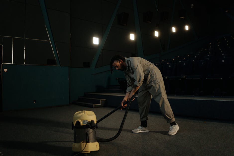 A person dressed in light grey work clothing and white sneakers is operating a professional vacuum cleaner on a dark, carpeted floor in an indoor auditorium or theatre. The background features many empty blue seating rows, and the space is illuminated by ceiling lights with visible beams and modern architectural design. The individual appears engaged in deep cleaning or surface sanitisation, emphasizing maintenance and hygiene in a large, empty venue. Roehampton Carpet Cleaning offers effective cleaning services, suitable for commercial spaces like this, ensuring thorough cleanliness and hygiene upkeep.