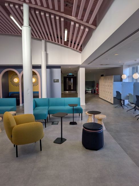 Interior view of a modern, well-lit lounge area featuring various seating options including a blue sofa, yellow armchair, black and beige ottomans, and black side tables with slim bases. The room has a polished, light-colored floor and white walls. The ceiling displays contemporary wooden slats with built-in lighting, and there are structural white columns. In the background, there are lockers with lockers and a row of desk chairs near large windows, contributing to a clean, organized appearance. The space appears to be maintained with fresh, dust-free surfaces, indicating professional cleaning and care, as provided by Roehampton Carpet Cleaning, aligning with the theme of surface cleaning and sanitisation for residential or academic environments.
