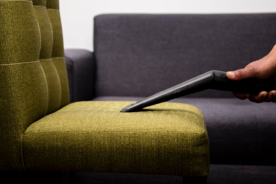 Close-up of a person's hand holding a vacuum cleaner nozzle, performing surface cleaning on a green upholstered armchair in a room with a dark gray sofa in the background. The armchair has a textured fabric with a tufted backrest and is situated on a neutral-colored floor. The lighting is even, highlighting the cleanliness of the fabric and the use of professional cleaning equipment by Roehampton Carpet Cleaning, as part of their upholstery cleaning and care services. The scene emphasizes a thorough domestic cleaning process focused on maintaining hygiene and upholstery appearance.
